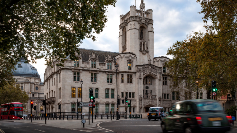 Supreme Court on a busy London street, framed by autumn trees; a red double-decker bus and cars pass by.
