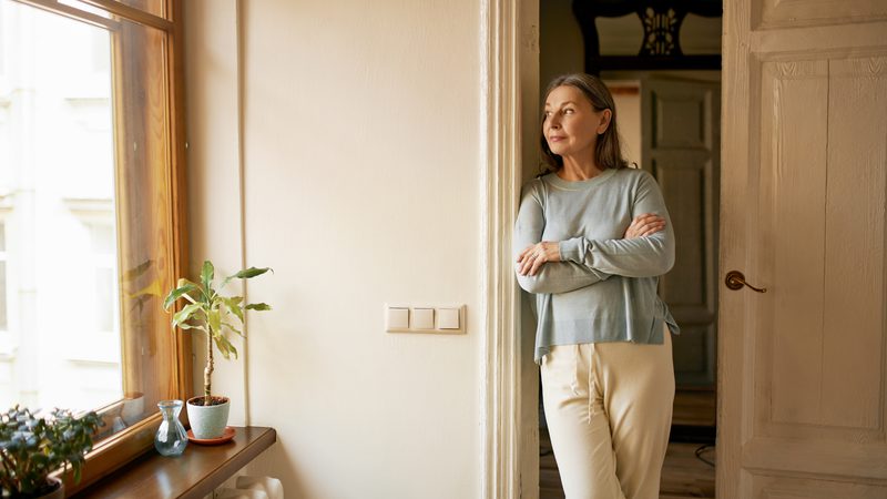 A woman with grey hair stands with arms crossed by a doorway, gazing thoughtfully out of a window. She wears a light grey top and cream trousers in a softly lit room.