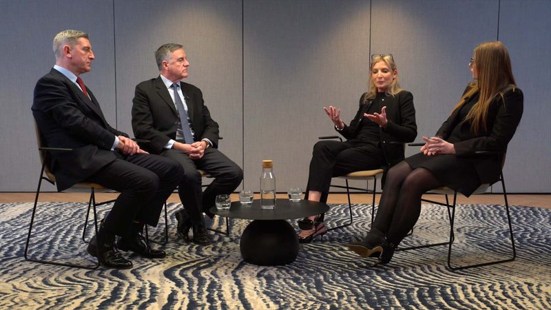 Four people in formal attire are seated in a semi-circle on chairs, engaging in a discussion; a woman in black is speaking while others listen attentively.