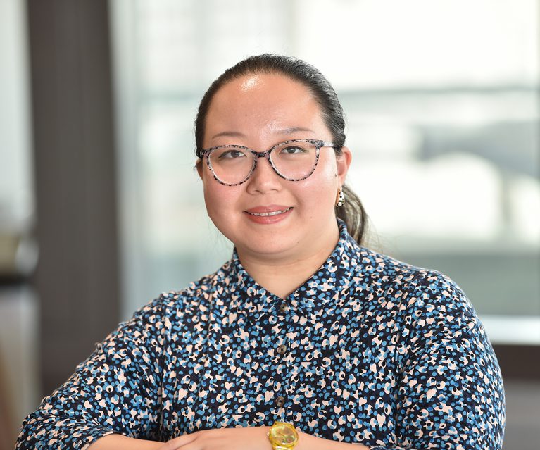 A woman with glasses and dark hair tied back, wearing a patterned blouse, sits indoors with a blurred background of windows and modern interior.