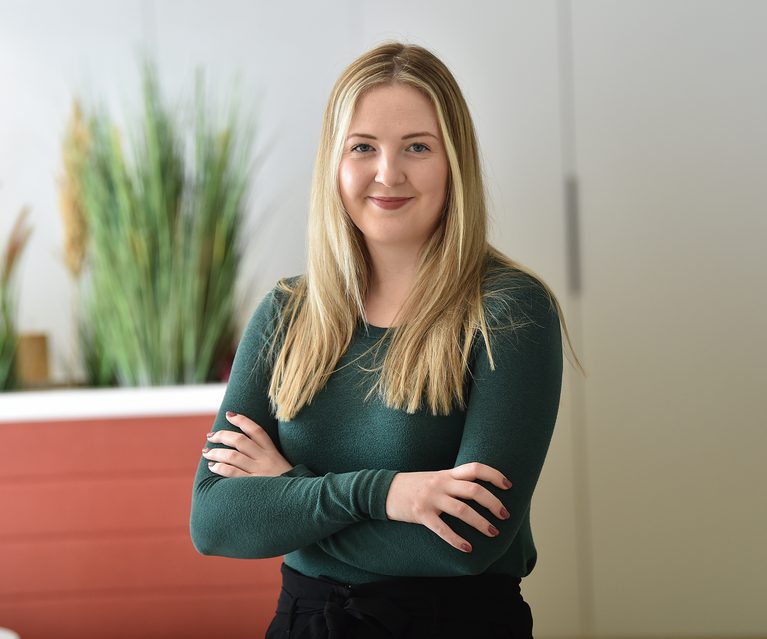 A young woman with long blonde hair and a gentle smile, wearing a dark green top, standing with arms crossed in a bright, modern office space.