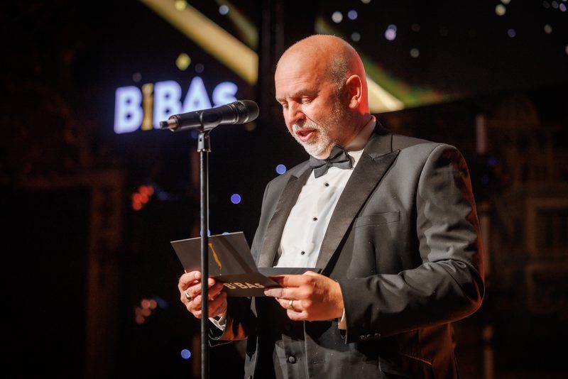 A person in a black tuxedo and bow tie speaks into a microphone while reading from a card on a dim stage, with blurred lights in the background.