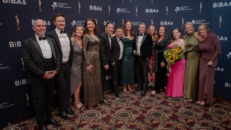 Group of formally dressed people posing for a photo at an awards event, standing in front of a blue backdrop with BiBAs logos and a patterned carpet.