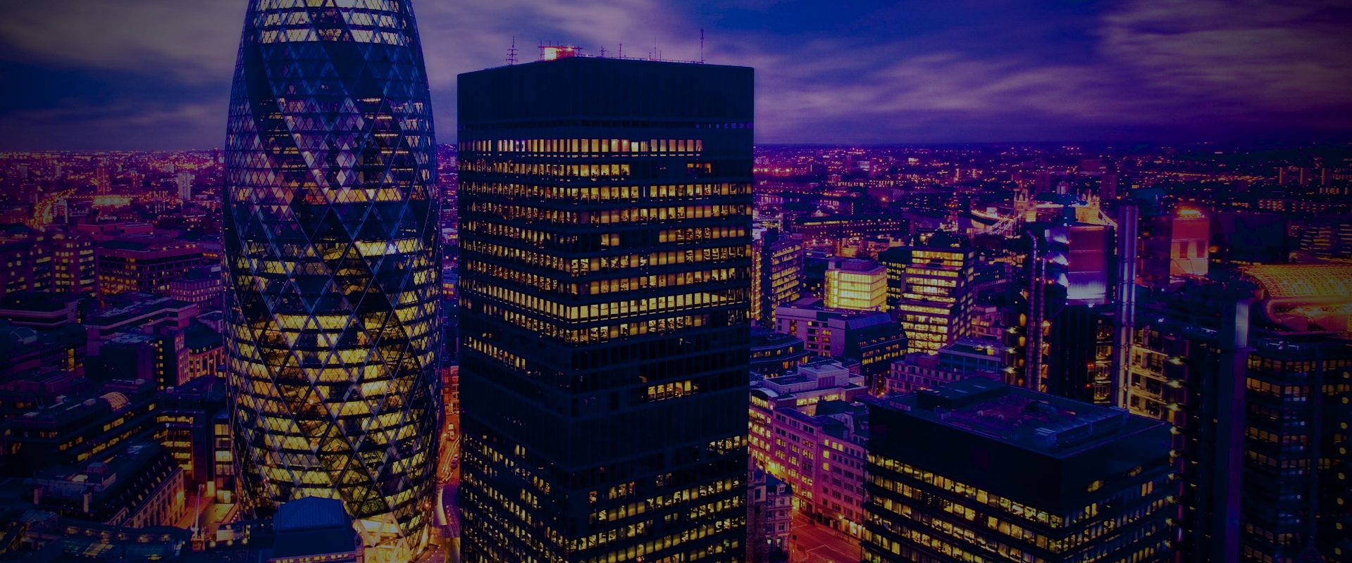 Night city skyline with lit windows on high-rise buildings and a distinctive diamond-patterned glass tower, beneath a purple-blue evening sky.