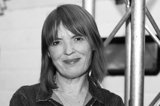 A woman with shoulder-length hair and a slight smile, wearing a denim jacket, standing indoors near metal scaffolding, in black and white.