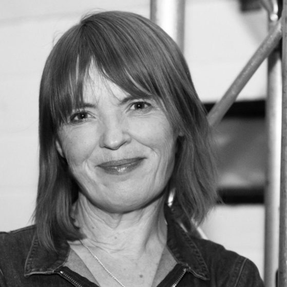 A woman with shoulder-length hair and a slight smile, wearing a denim jacket, standing indoors near metal scaffolding, in black and white.