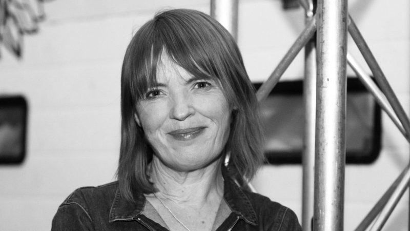 A woman with shoulder-length hair and a slight smile, wearing a denim jacket, standing indoors near metal scaffolding, in black and white.