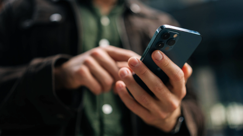 Person holding and using a smartphone outdoors, wearing a dark jacket and green shirt.