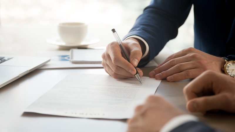 A person in a dark suit is signing a document on a desk with a pen, with a coffee cup, holder, and a laptop nearby.