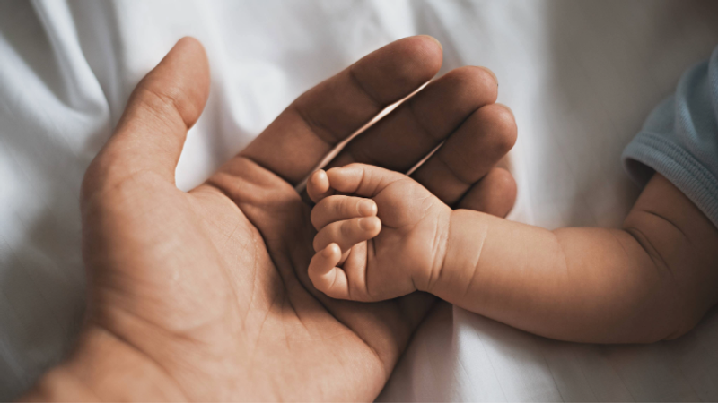 Close-up of an adult hand gently cradling a tiny baby's hand, fingers interlaced in a tender grip, resting on a white bedsheet backdrop bathed in warm light.
