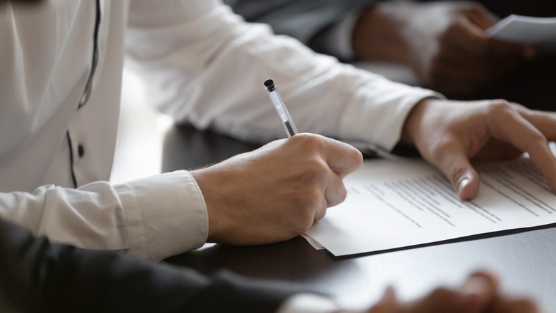 A person in a white shirt is writing on a document with a pen, with other people visible in the background.