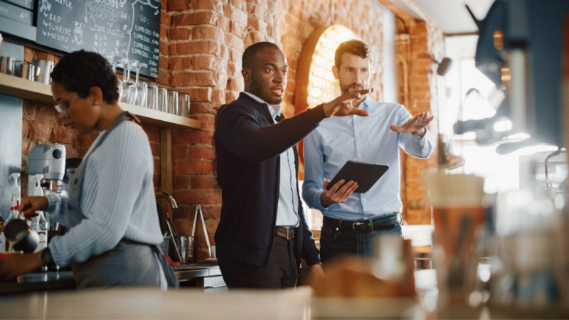 Two men in business attire are having a discussion behind the counter of a coffee shop, with one holding a tablet and gesturing as they talk. A woman operates a coffee machine nearby.
