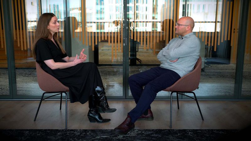 A woman and a man sit facing each other in chairs, engaged in a conversation in a modern office space with glass walls and wooden flooring.