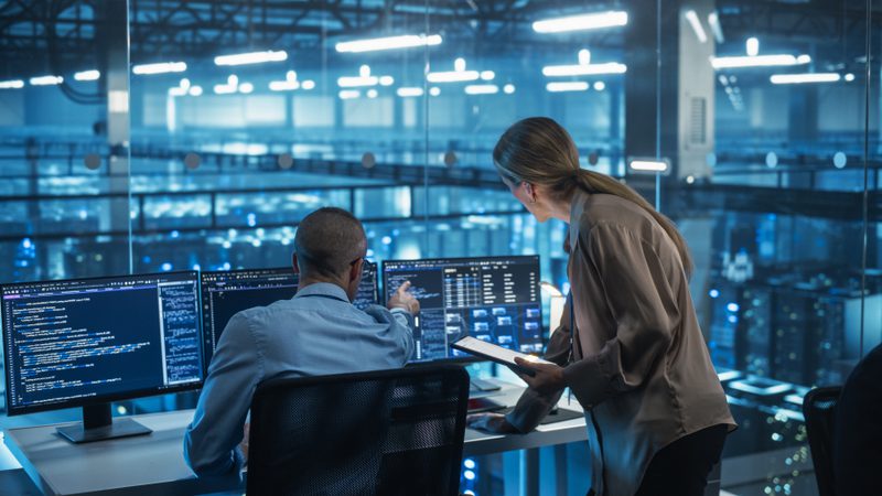 Two professionals are working in a high-tech environment with multiple computer screens displaying code and data. One person is seated at a desk, and the other is standing, consulting a tablet.