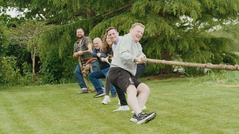 A group of people outdoors on a grassy lawn pull a thick rope in a tug-of-war, smiling and leaning back against a backdrop of tall, leafy trees.