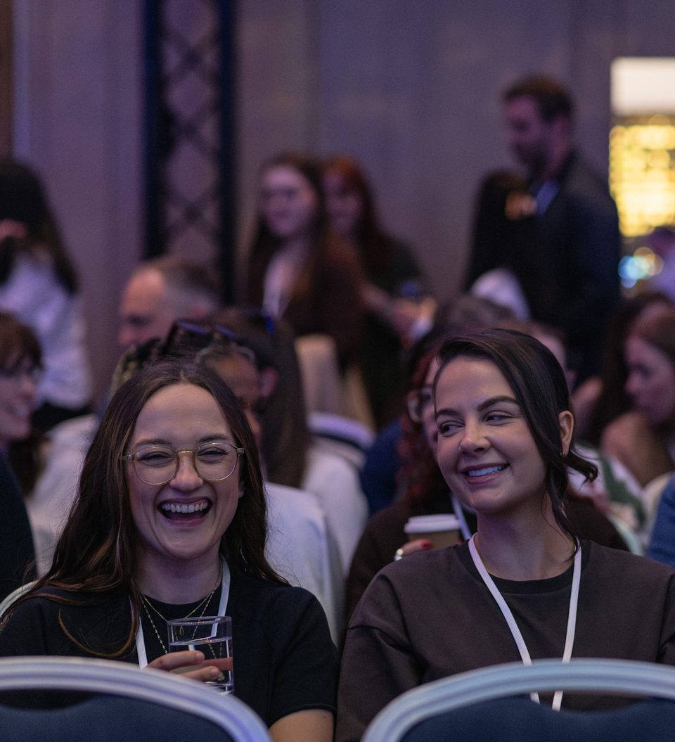Three attendees in the foreground laughing and chatting at a crowded indoor event; one holds a glass while others smile, with blurred guests and warm backlighting in the background.