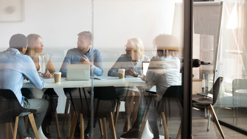 People sit around a glass-walled conference room table with laptops and coffee, as a flip chart stands nearby in a bright office.