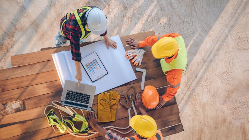 Three construction workers wearing helmets and safety gear are gathered around a wooden table, reviewing plans and discussing tools on a sandy surface.