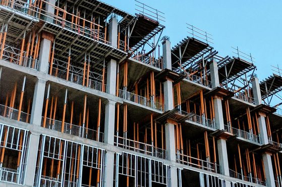 A multi-storey building under construction with exposed concrete, orange scaffolding, and metal framework against a blue sky.