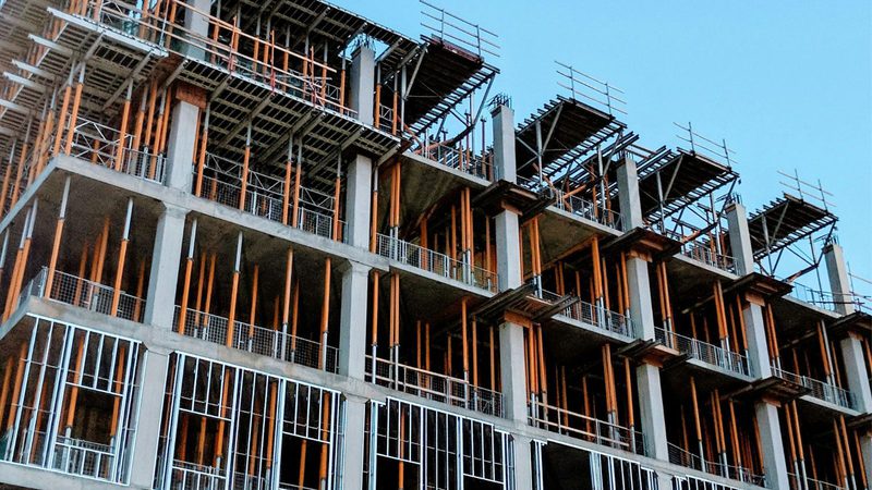 A multi-storey building under construction with exposed concrete, orange scaffolding, and metal framework against a blue sky.