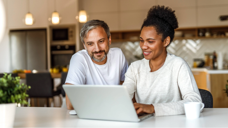 A man and a woman sit together at a table, looking at a laptop screen in a modern kitchen. They are smiling and appear engaged with what they are viewing.