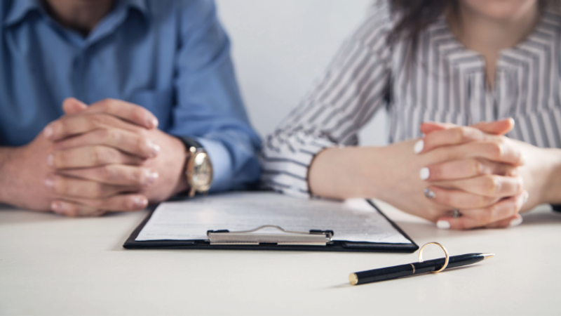 Two people sit at a table with a clipboard and a pen, hands clasped in front of documents, suggesting a meeting or agreement.