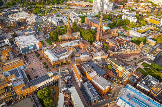 Aerial view of a city centre with historic churches, modern buildings, and green parks, showing a mix of architecture, roads, and open plazas.