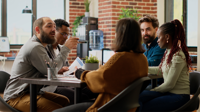Five diverse colleagues sit around a table in a bright office, discussing documents and a laptop, with a plant and water cooler against a brick wall.