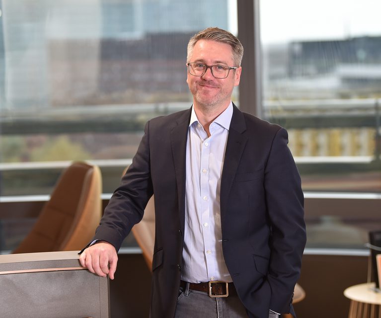 A professionally dressed man with glasses and a beard stands in an office, leaning on a desk with a window backdrop showing a cityscape.