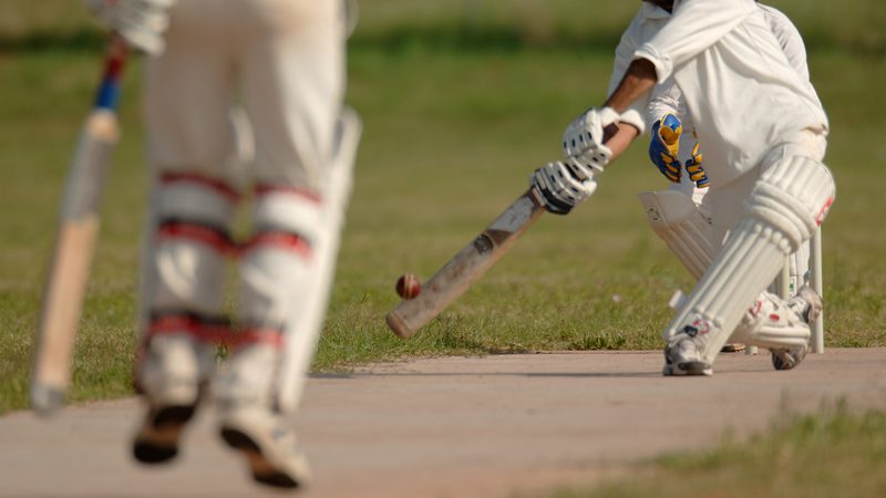 A cricket player in white pads and gloves is attempting a shot with a cricket bat, while another player in white protective gear watches on the grassy field.