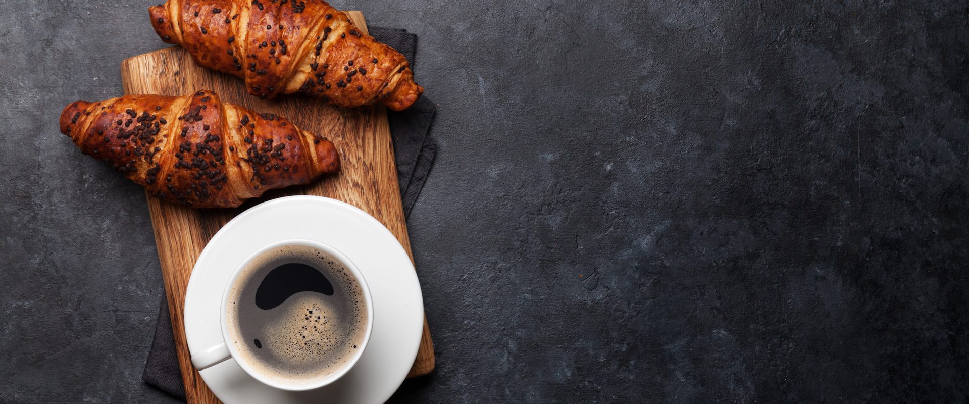 Two croissants topped with chocolate chips on a wooden board, alongside a cup of black coffee on a white saucer, placed on a dark textured surface.