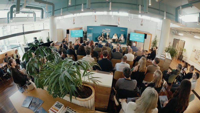 Panel discussion in a bright modern conference room with a blue backdrop reading Making Places Work, audience facing the stage, plants in the foreground.