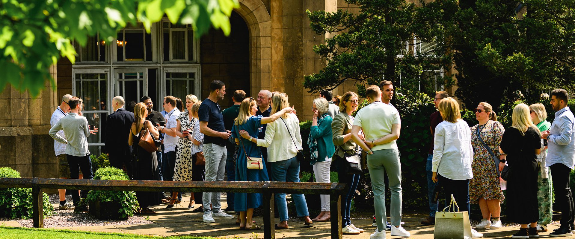 Group of people standing and socialising outdoors on a sunny day near a historic building, with green trees and grass in the background.
