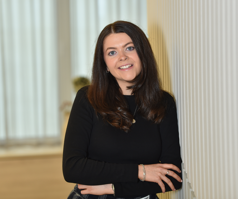A woman with long, dark brown hair and fair skin, wearing a black long-sleeve top, standing indoors against a light striped wall, smiling at the camera.