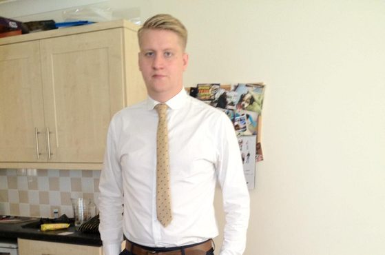 A young man with blonde hair, wearing a white shirt, beige tie, dark trousers, and brown shoes, stands indoors with hands in pockets near a kitchen area.