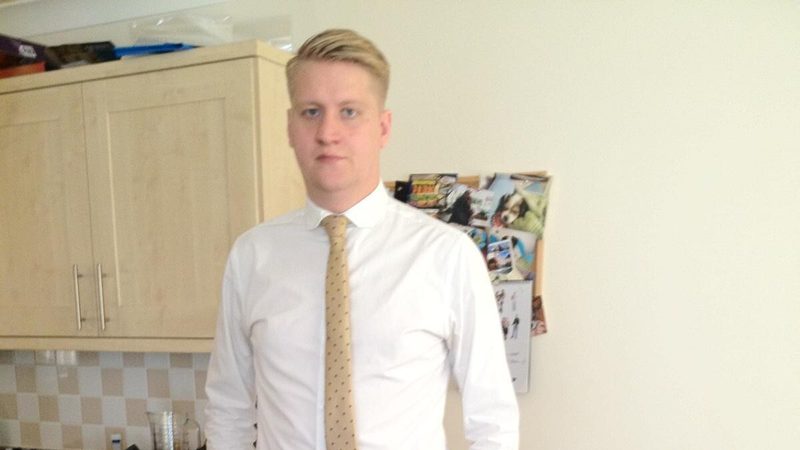 A young man with blonde hair, wearing a white shirt, beige tie, dark trousers, and brown shoes, stands indoors with hands in pockets near a kitchen area.