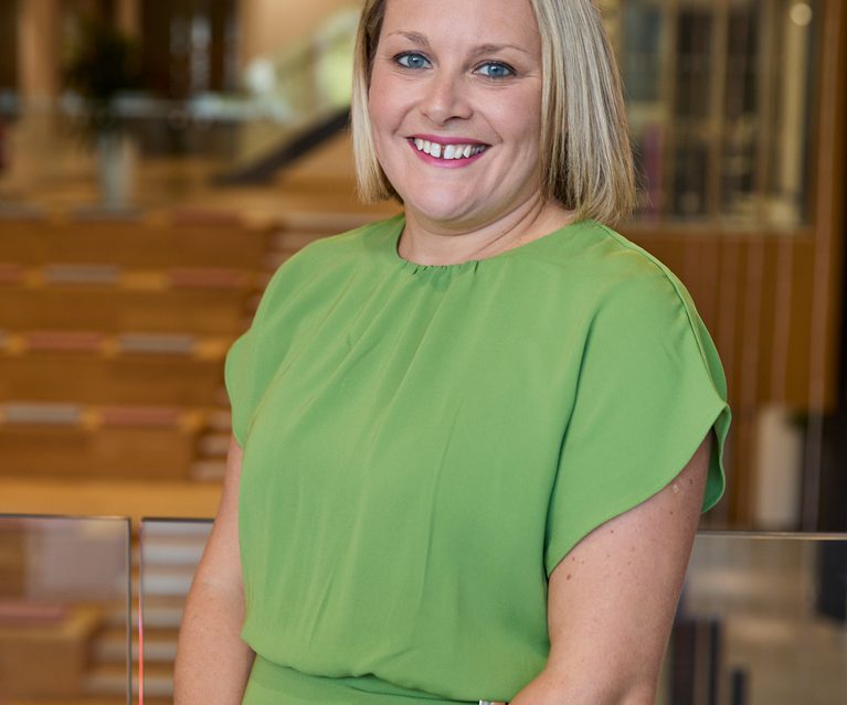 Smiling person with shoulder-length blonde hair wearing a lime-green dress stands by a glass railing in a modern office lobby with warm lighting and wood seating.