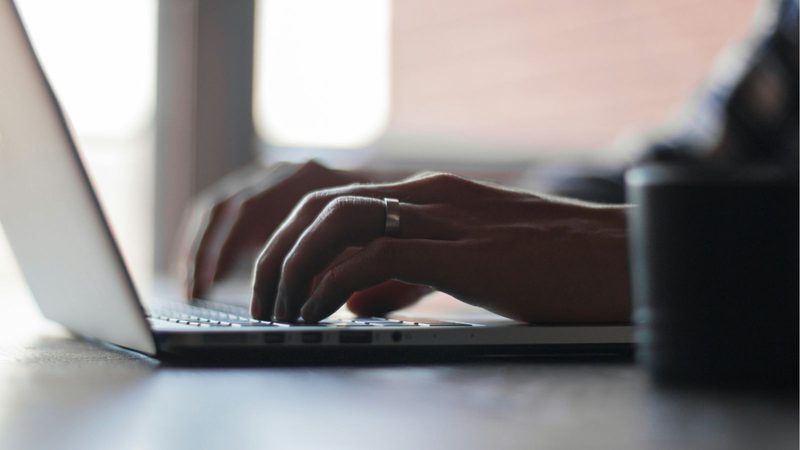 Hands typing on a laptop keyboard with a window in the background, softly illuminated and slightly out of focus.