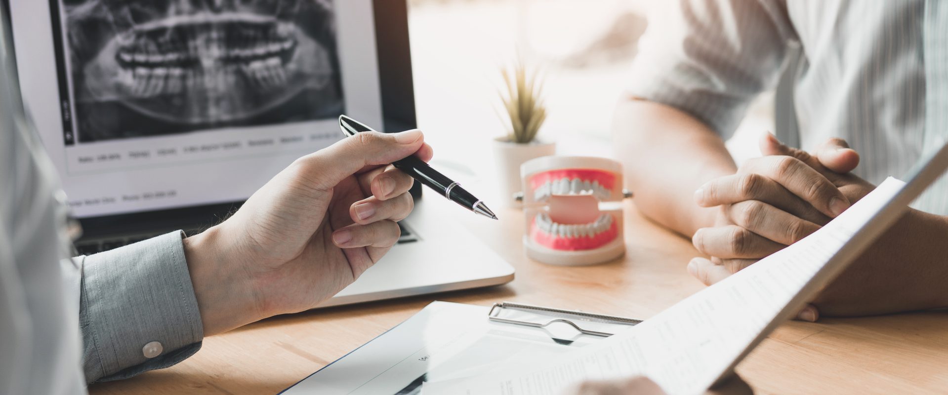 A person holds a pen while showing dental x-rays and a dental model, discussing oral health in a consultation with another individual.