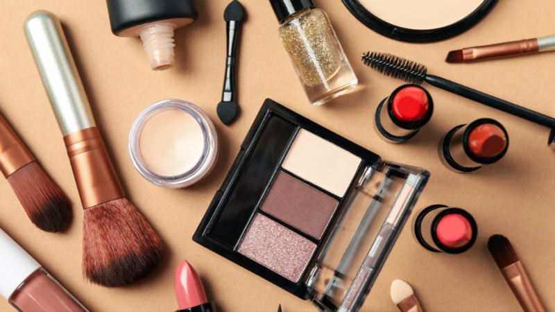 Assorted makeup items spread across a warm beige background, including brushes, lipsticks, nail polish, an eyeshadow palette, compact powder, and various beauty tools, artfully arranged.