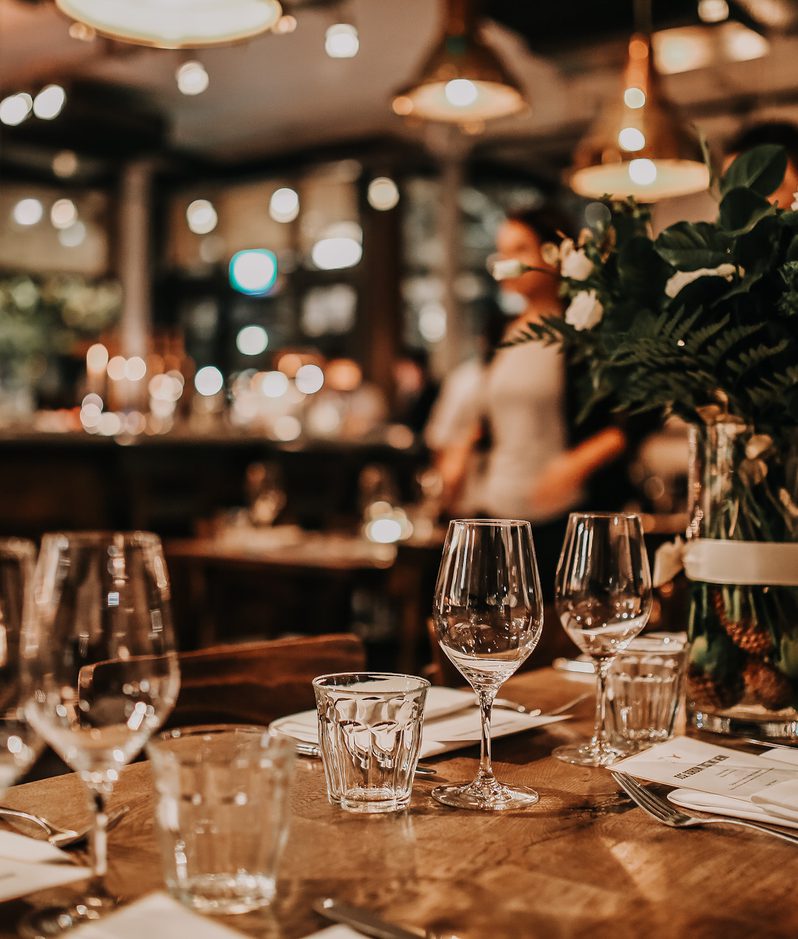 A warmly lit restaurant table set with empty wine glasses, water glasses, and white napkins, with a large floral arrangement in the background.