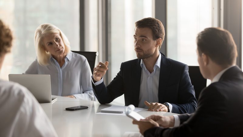 A group of four professionals engaged in a discussion around a white table in a modern office with large windows.