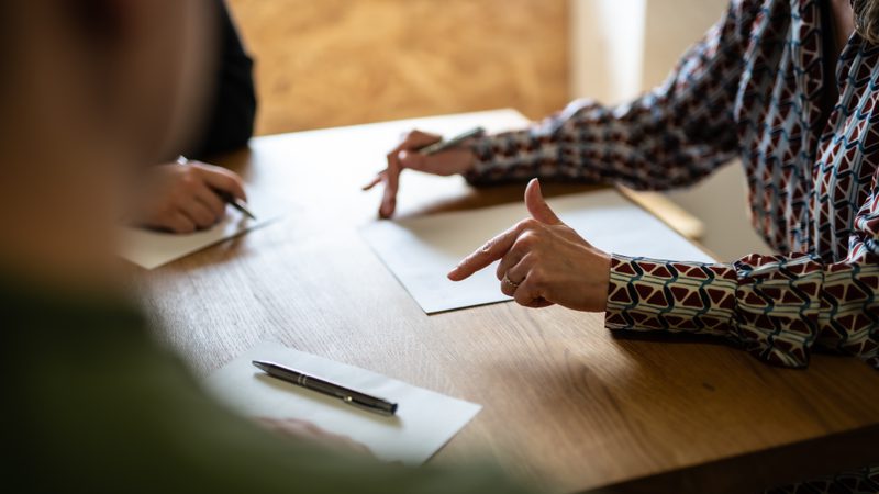 A person in a patterned shirt points at a paper on a wooden table during a discussion, with other people also engaged, writing on notepads.