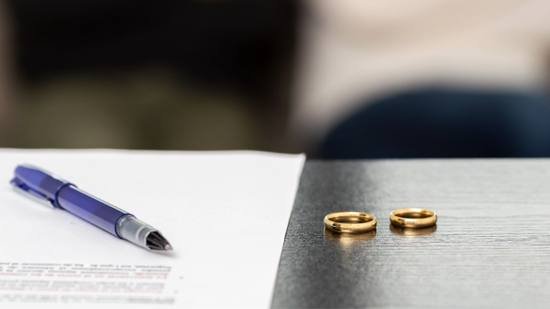 Blue ballpoint pen rests on a white sheet of paper, while two gold wedding rings lie on a dark wooden desk; a blurred backdrop softens the scene.