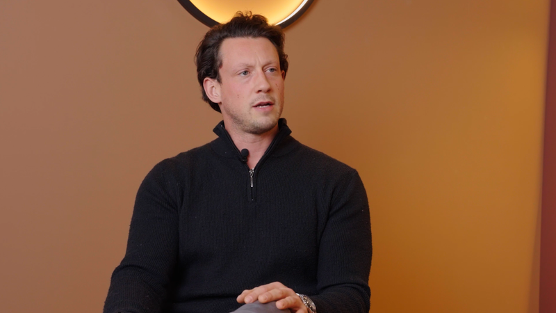 A man with dark, wavy hair wearing a black quarter-zip sweater sits against a warm orange background, gazing to the left with a thoughtful expression.