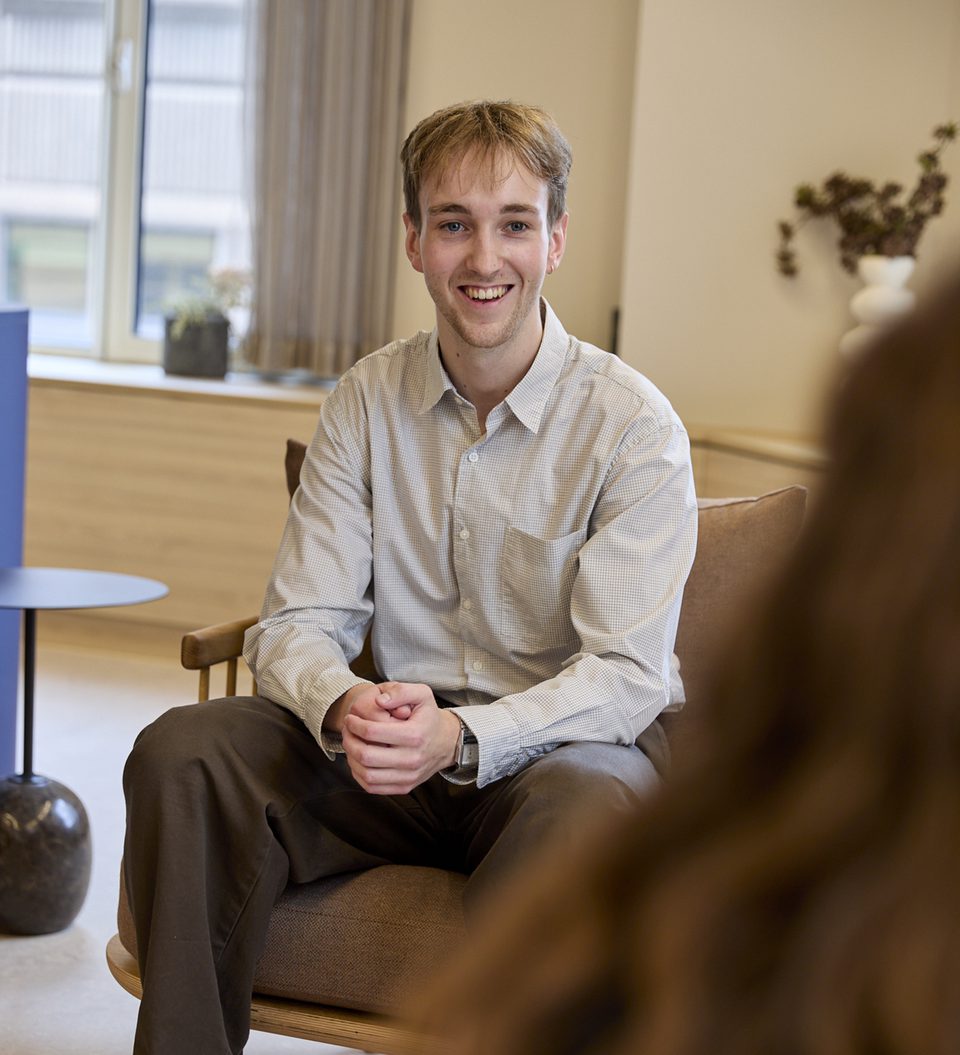 A person sits on a beige sofa, smiling, wearing a light check shirt and brown trousers, in a bright room with a window and potted plants.