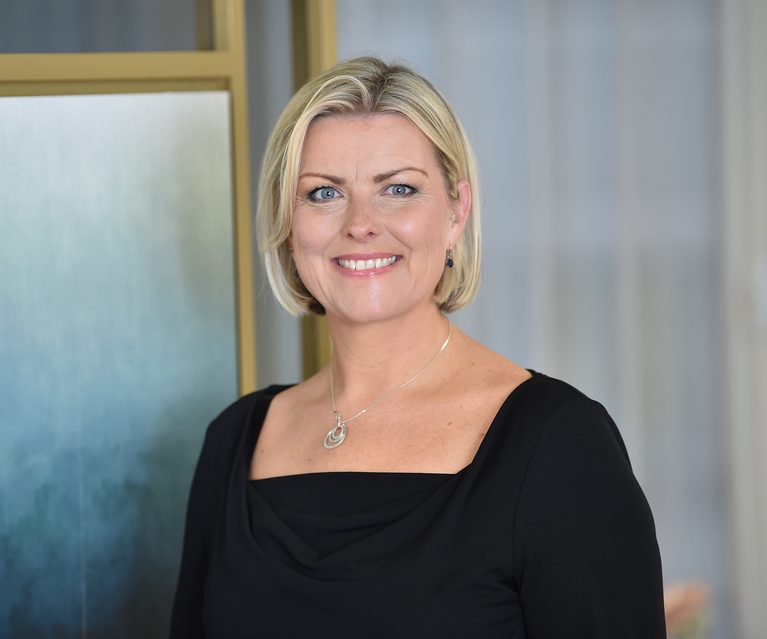A woman with short blonde hair, blue eyes, and fair skin, smiling and wearing a black top with a silver necklace, standing indoors with a partly blurred background.