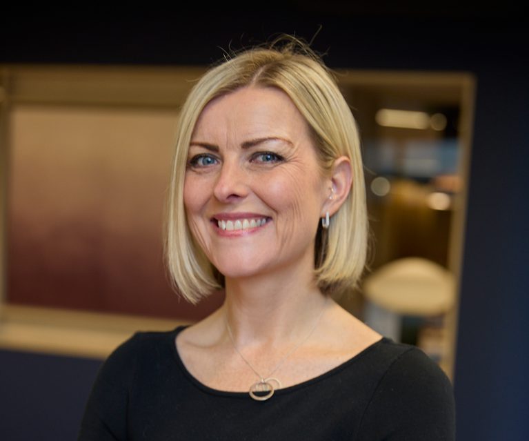 A woman with short blonde hair, blue eyes, and fair skin, smiling and wearing a black top with a silver necklace, standing indoors with a partly blurred background.