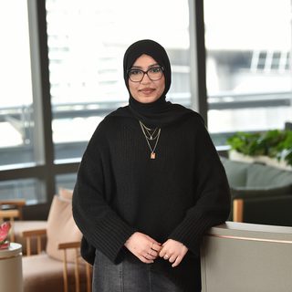 A woman in a black hijab, glasses, and a black sweater standing in a modern office space with large windows, greenery, and comfortable seating.