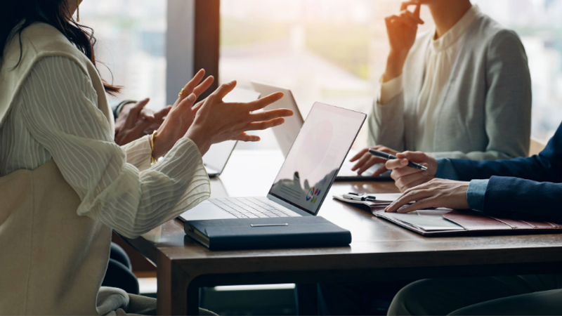 Three people in a meeting, engaging in discussion at a table with laptops and documents, with natural light coming through a large window in the background.